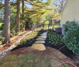natural stone path wrapping around a house surrounded by flowerbeds of new growth with drainage off to the side to prevent flooding
